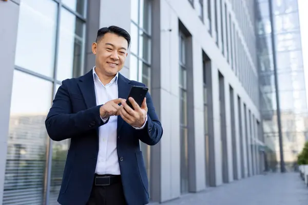 A smiling young Asian male businessman in a suit is standing outside an office center and using a mobile phone.