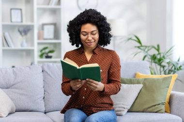 Evdeki kanepede oturan, gözlüklü ve konsantre bir kitap okuyan Afro-Amerikalı genç bir kadın..