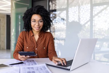 Ofiste çalışan genç bir Afro-Amerikalı kadının portresi. Bilgisayarlı, belgeli ve faturalı, elinde bir hesap makinesi olan ve kameraya gülümseyen..