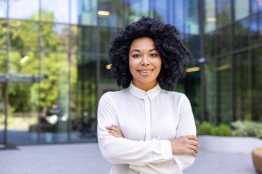 Beyaz gömlekli, gülümseyen ve başarılı bir iş kadınının yakın plan portresi. Kollarını göğsünün üzerinden geçirip kendine güvenle kameraya bakıyor..