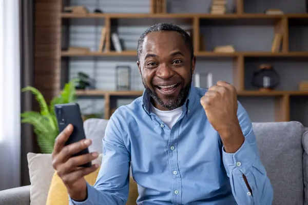Portrait of a happy African American man sitting on the couch at home, using a mobile phone, and looking happy at the camera showing a victory gesture with his hand.