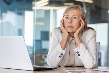 Sad unsatisfied senior woman at workplace depressed, mature business woman unhappy with achievement results at workplace unhappy sitting at table with laptop, boss thinking about problems. clipart