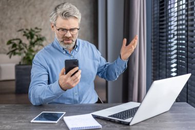 Sad disappointed boss businessman at workplace received online message email on phone, senior mature gray haired man using app on smartphone, inside office with laptop. clipart