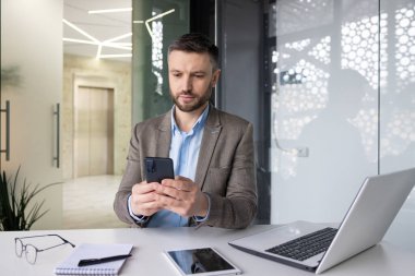 Çevrimiçi sosyal ağlar okumayı düşünen ciddi konsantre iş adamı, iş yerinde bilgisayarlı akıllı telefon kullanan deneyimli yetişkin bir adam..