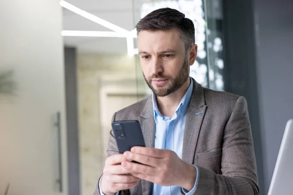 Serious concentrated mature businessman thinking reading online social networks, experienced mature man holding phone using smartphone at workplace inside office with laptop.