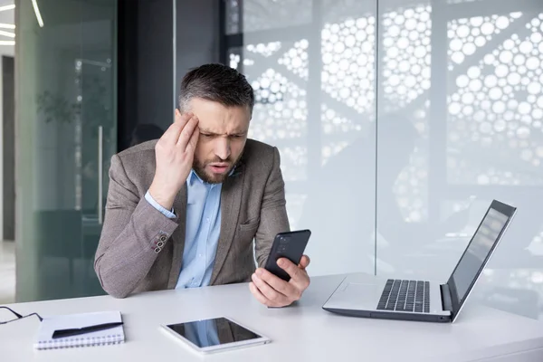 Upset frustrated man holding phone inside office, sad businessman reading bad news online, boss experienced financier accountant unhappy with workplace achievement results.