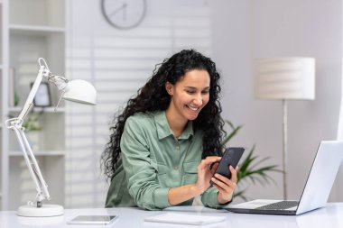 İş kadını iş yerinde mutlu, telefon kullanıyor, Latin Amerikalı mutlu akıllı telefondaki uygulamayı kullanıyor, sosyal ağlara takılıyor, ev ofisinde bilgisayarla oturuyor..