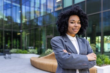 Gülümseyen ve kendinden emin bir Afrikalı Amerikalı iş kadınının portresi ofis binasının önünde duruyor ve kollarını göğsüne bağlamış kameraya bakıyor..