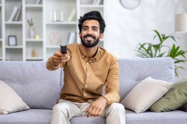 Gülümseyen bir Hintli genç adam evde dinleniyor, kanepede oturuyor ve uzaktan kumandayla TV 'yi açıyor..
