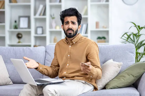 Portrait of a worried young Indian man sitting on the sofa at home, holding a credit card and using a notebook, looking at the camera in frustration and throwing up his hands.