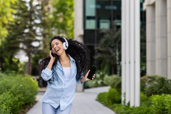 Young joyful beautiful woman dancing and singing in headphones while walking in the city, Hispanic woman with curly hair uses an application on the phone, to listen to music online.