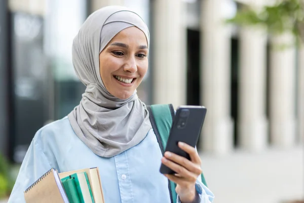 Young muslim woman in hijab walking outside university campus, female student smiling contentedly using app on phone, backpack on back and books in hands.