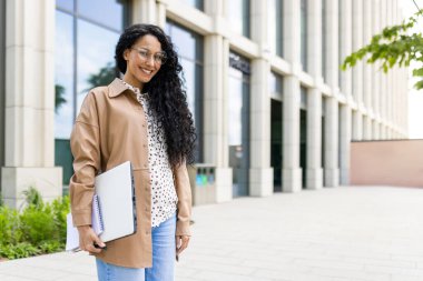 Ofis binası dışında başarılı bir iş kadını portresi, başarılı bir Latin Amerikalı kadın gülümseyen ve kameraya bakan, dizüstü bilgisayar ve belgelerle ayakta duran.