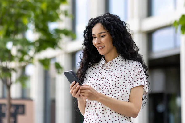 A young beautiful Latin American woman with curly hair walks through the city with a phone in her hands, a woman uses an application on a smartphone, browses Internet pages and social networks.