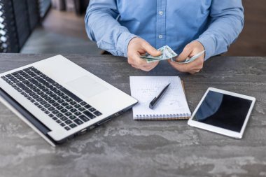 Close-up photo of the hands of a man sitting at a desk in the office and counting money. At workplace with laptop, tablet and notepad.