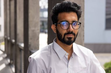 Portrait of a close young businessman, outside the office building, a man is confidently looking in the camera, a worker in the shirt is thinking and focused.