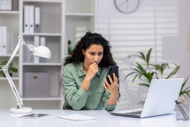 Ofiste oturan, üzgün, genç bir İspanyol kadın telefon ekranına endişeli bir şekilde bakıyor..