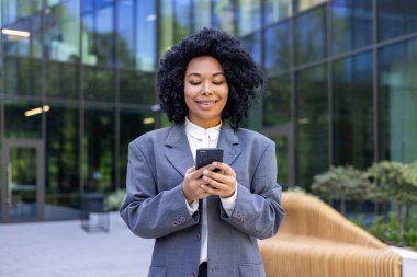 Başarılı bir Afro-Amerikan iş kadını ofis binası dışından şehre geliyor, finans çalışanı mutlu bir şekilde gülümsüyor, akıllı telefonu elinde tutan kadın uygulamayı kullanıyor, sosyal ağları tarıyor..