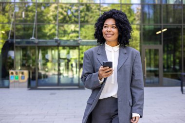 Başarılı bir Afro-Amerikan iş kadını ofis binası dışından şehre geliyor, finans çalışanı mutlu bir şekilde gülümsüyor, akıllı telefonu elinde tutan kadın uygulamayı kullanıyor, sosyal ağları tarıyor..