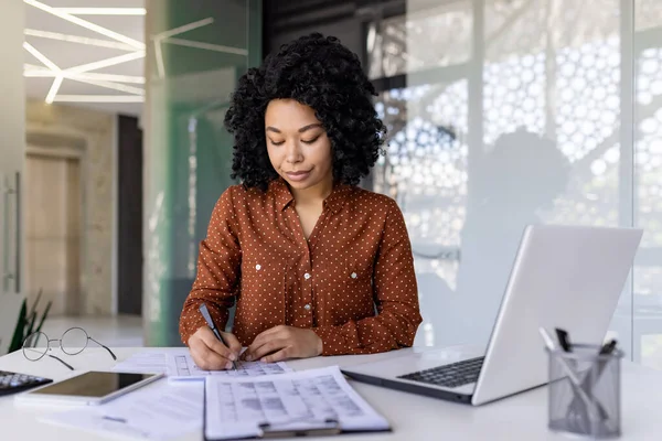 Mature experienced business woman filling forms and tables while sitting at workplace inside office, financial accountant serious and concentrated at work, recording data in reports.
