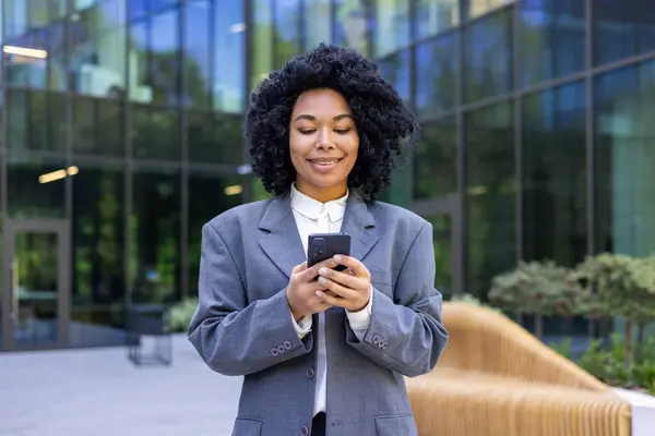 Successful african american business woman walking in the city from outside office building, financial worker smiling contentedly, woman holding smartphone using app, browsing social networks.