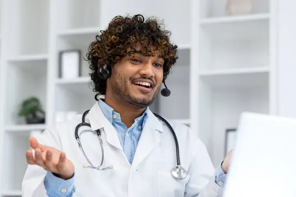 Young Indian doctor consulting patients remotely, man in white medical coat with headset using laptop for video call, smiling contentedly inside clinic office.
