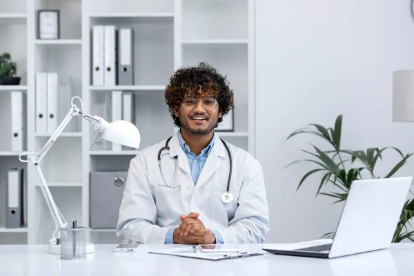 Portrait of young Indian doctor, man in white medical coat smiling and looking at camera, doctor sitting at table inside medical office of clinic, working with laptop.