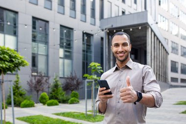 Gömlekli bir adam modern bir ofis binasının önünde elinde cep telefonuyla baş parmak işareti yapıyor.