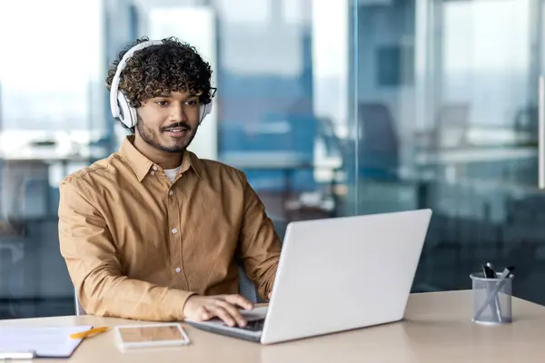 Young indian programmer working inside office with laptop, man in headphones coding new software, businessman developer satisfied with achieved results smiling.