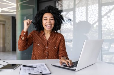 Ofiste dizüstü bilgisayarla oturan ve kameraya mutlu bir şekilde bakan Afro-Amerikalı bir iş kadınının portresi, bir başarı ve zaferin el hareketini gösteriyor..