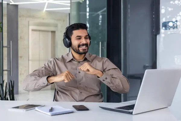 A young Indian man works in the office. He sits at his desk with headphones in front of his laptop and dances during the break, rests, rejoices.