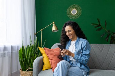 Oturma odasındaki kanepede oturan güzel İspanyol kadın mutlu bir şekilde gülümsüyor telefonu elinde tutuyor, akıllı telefondaki uygulamaları kullanıyor, interneti ve sosyal ağları karıştırıyor..