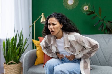 Sağlıksız kadın evdeki kanepede oturuyor, İspanyol kadın ağır mide ağrısı çekiyor, el ele tutuşuyor..