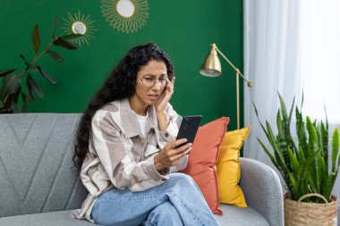 Üzgün, mutsuz bir kadın evdeki oturma odasında oturuyor, İspanyol bir kadın elinde telefonla, mutsuz bir şekilde mesajlar okuyor ve sosyal haberleri kontrol ediyor..