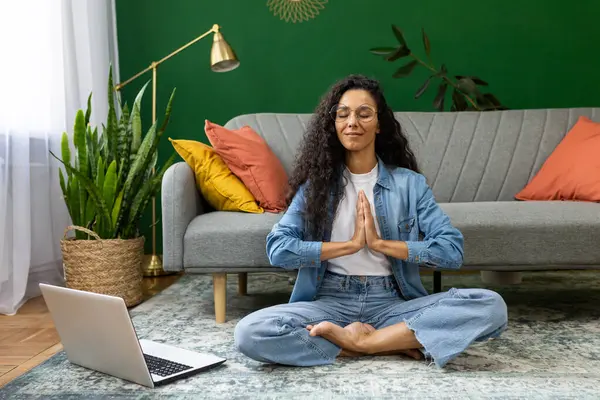 Nilüfer çiçeği pozunda dinlenen genç bir kadın oturma odasında yerde oturuyor dizüstü bilgisayarlı İspanyol kadın eğitim sınıfında internetten yoga yapıyor..