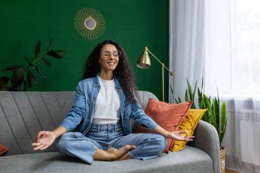 Nilüfer çiçeği pozunda meditasyon yapan genç, güzel bir kadın, İspanyol kadın mutlu bir şekilde gülümseyen, gelecekle ilgili planlar kuran, evdeki oturma odasında kanepede oturan, yoga kursuna çalışan,.
