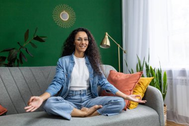 Nilüfer çiçeği pozunda meditasyon yapan genç, güzel bir kadın, mutlu bir şekilde gülümseyen ve kameraya bakan İspanyol bir kadın, evdeki oturma odasında kanepede oturup yoga dersleri alan bir kadın..
