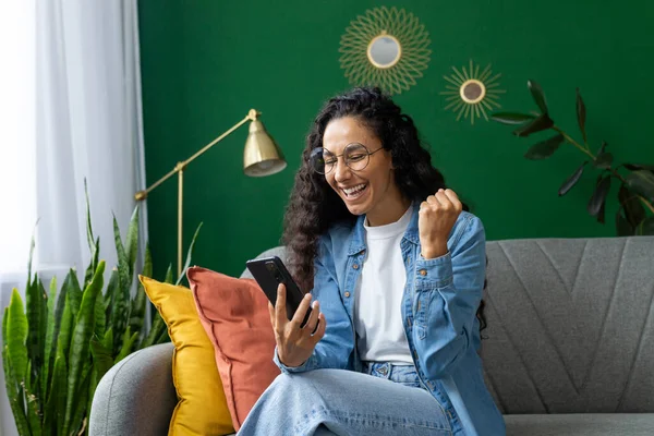 A young woman is pictured indoors, showing a burst of joy and excitement as she looks at her smartphone, possibly celebrating a piece of good news or a personal achievement.