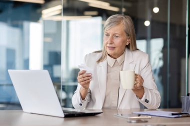 Ofiste çalışan hasta kıdemli bir iş kadını, masada oturuyor, elinde bir bardak ve peçeteyle, endişeli bir şekilde dizüstü bilgisayarına bakıyor..