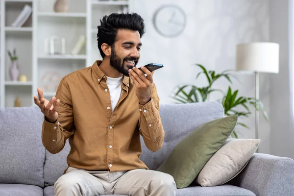 Joyful man speaking into smartphone like a microphone on a cozy sofa, expressing happiness and comfort in a modern living room.