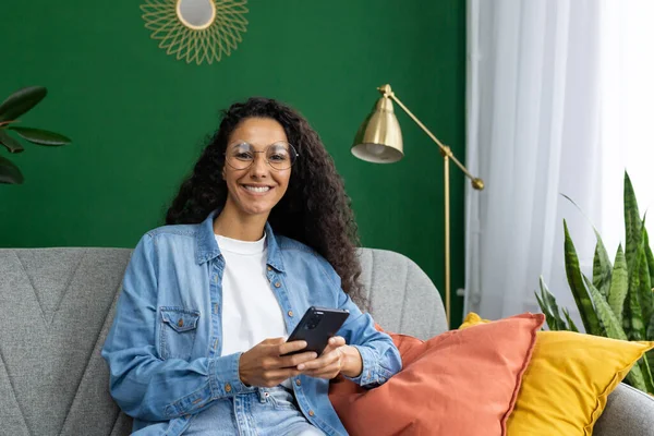 Smiling woman with glasses sitting on couch at home, holding phone. Cozy, stylish interior with green wall and plants.