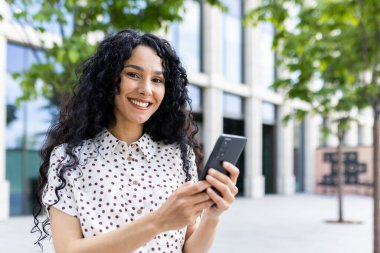 Kıvırcık saçlı neşeli kadın, şehir ortamında akıllı telefon kullanıyor, pozitiflik ve modern yaşam tarzı yayıyor..