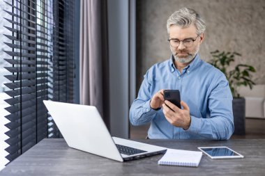 Masasında dizüstü bilgisayarı ve tableti olan akıllı telefon kullanan, odaklanmış ve ciddi iş kavramı olan olgun bir işadamı.