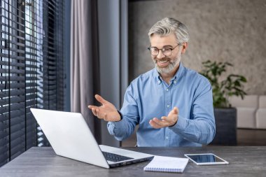 Mavi tişörtlü, gülümseyen olgun bir adam modern ofis ortamında dizüstü bilgisayarında bir video görüşmesi yapıyor ve pozitifliğini ifade ediyor..
