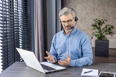 Kulaklık takmış olgun bir adam dizüstü bilgisayarda konuşuyor kendinden emin bir çalışan, online danışma, modern ofis geçmişi..