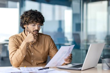 Düşünceli, profesyonel bir erkek derin odaklı belgeleri gözden geçirir. Bilgisayarlı ofis ayarları ve kağıtlar bir analiz anını yakalıyor.