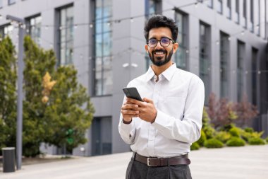Sakallı, kendine güvenen genç bir iş adamı. Açık havada akıllı telefon kullanırken gülümsüyor. Ofis arkaplanı..