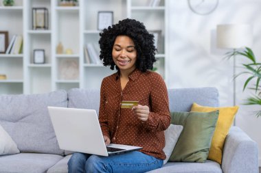 Genç Afrikalı Amerikalı bir kadın elinde kredi kartı ve dizüstü bilgisayarla kanepede oturuyor. İnternetten sipariş veriyor, alışveriş yapıyor, faturaları ödüyor..
