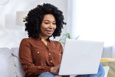 Evdeki kanepede oturan ve dizüstü bilgisayar kullanan gülümseyen Afro-Amerikan bir kadının yakın plan fotoğrafı..