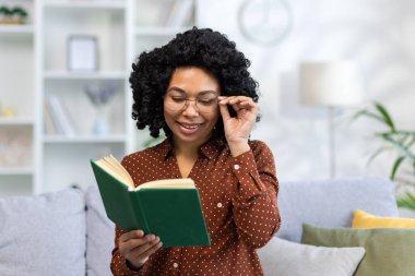 Konsantre olmuş gülümseyen Afro-Amerikan kadın evinde kanepede oturuyor ve ilginç bir kitap okuyor, elinde bardak tutuyor, boş zamanlarını geçiriyor, ders çalışıyor..